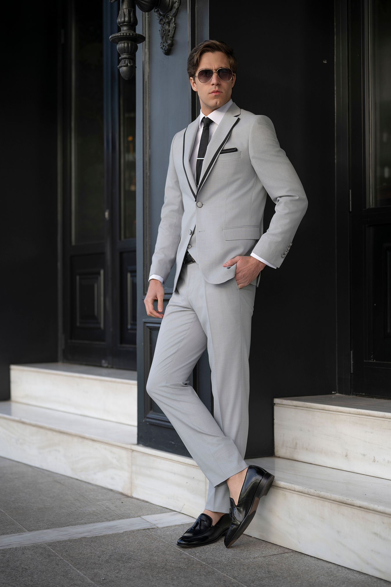 Σύγχρονο γαμπριάτικο look σε γκρι κοστούμι, σε στυλάτη στάση με loafers και γυαλιά.
